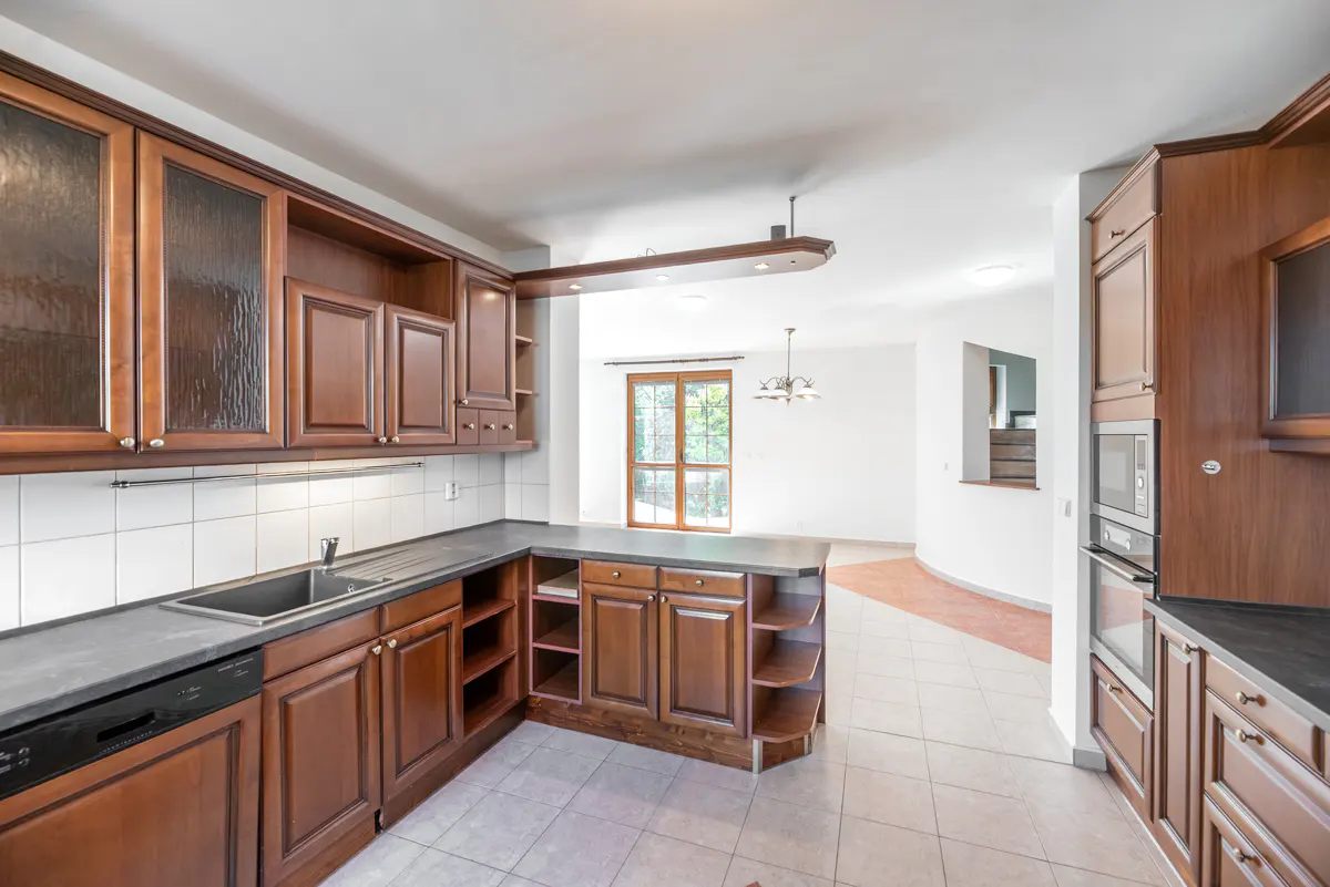 A kitchen with dark wood cabinets, a gray countertop, and white tile floors. A doorway leads to a bright, white room.