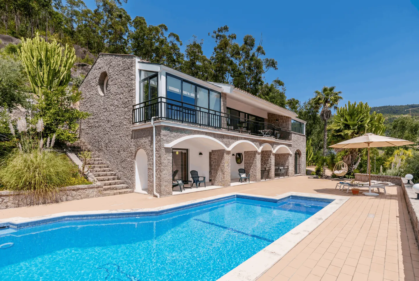 Exterior view of a stone house with a blue pool, patio furniture, and green trees under a clear blue sky.