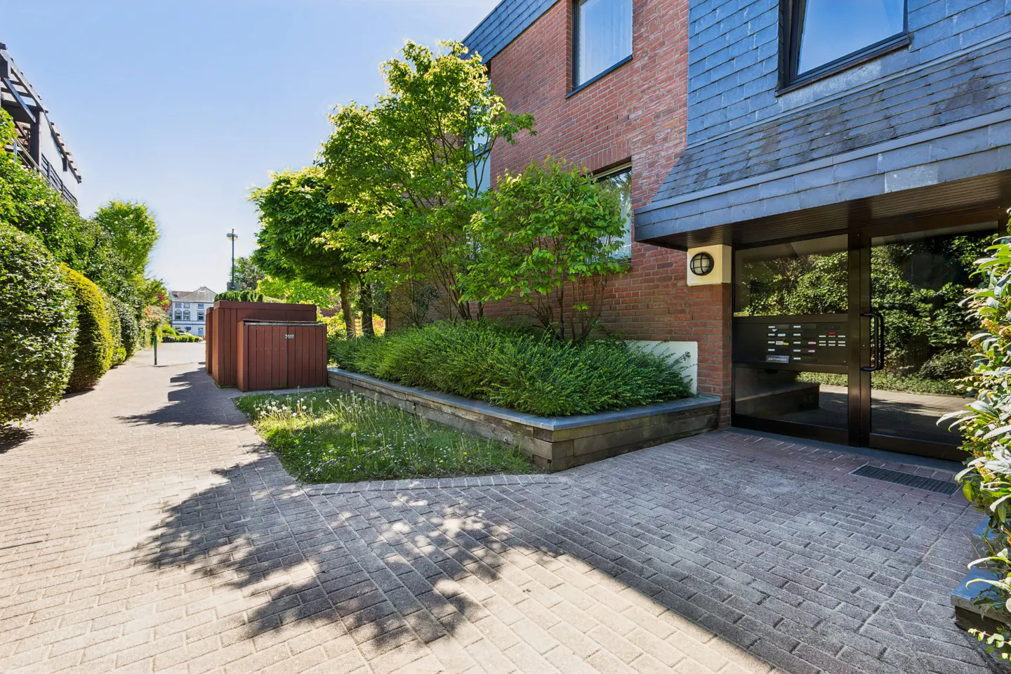 Apartment building entrance with brick facade, slate roof, and glass doors. Lush green landscaping and a brick walkway lead to the entrance.