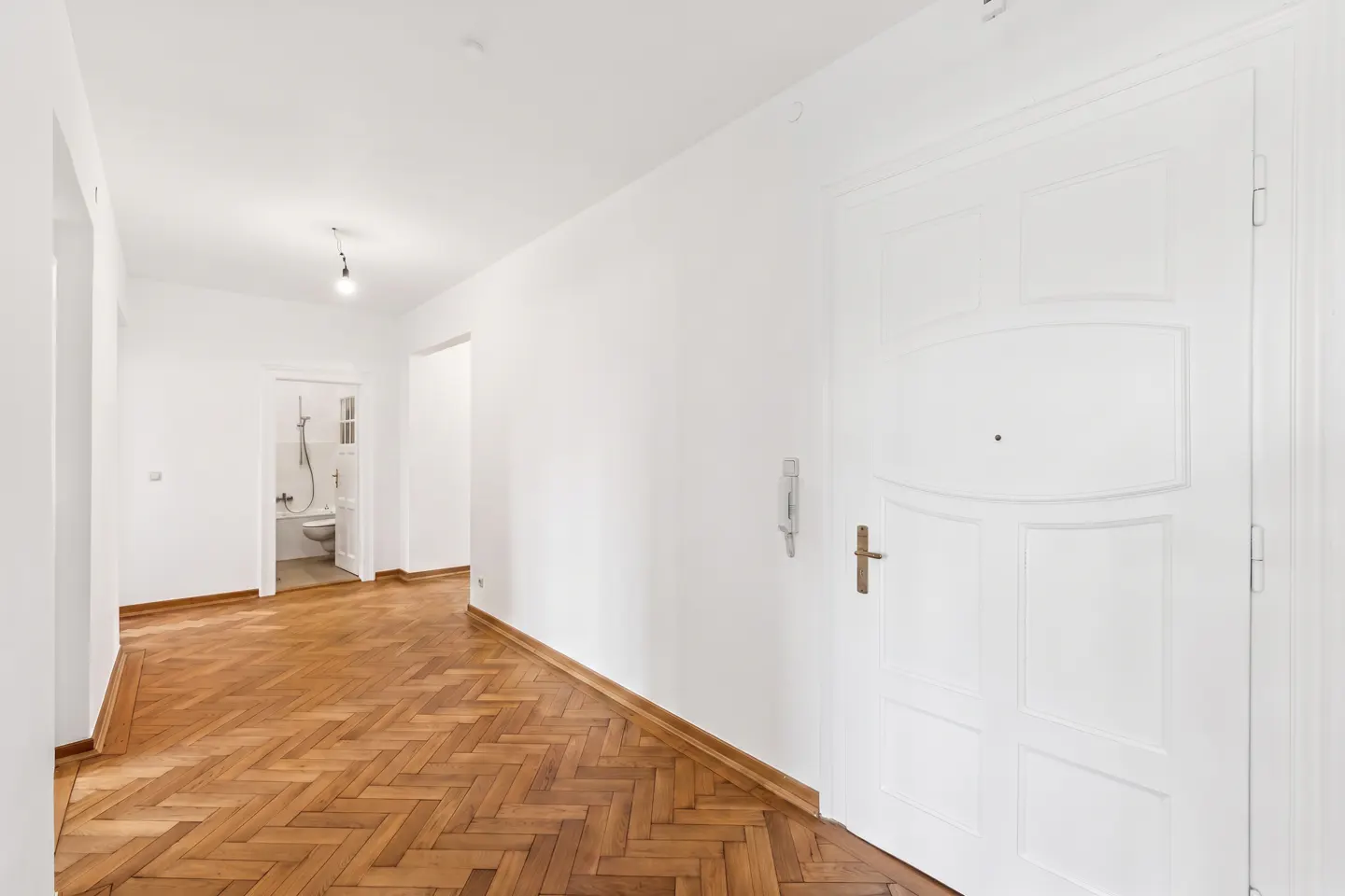 Bright, empty hallway with herringbone wood floors and white walls. A bathroom is visible in the distance. A white door is on the right.