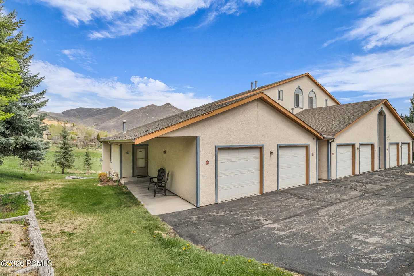 Exterior view of a beige townhouse with white garage doors, a green lawn, and mountains in the background under a blue sky.