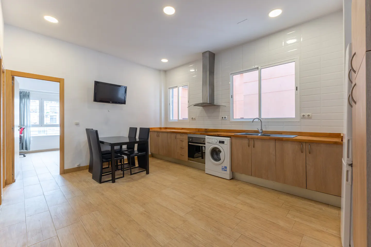 Bright, modern kitchen with wood floors, cabinets, and countertops. A black dining table sits near a wall-mounted TV. A washing machine is next to the sink.
