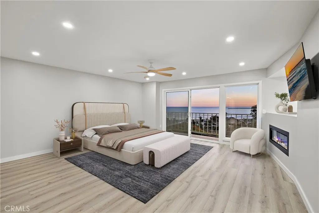 Bedroom with a bed, bench, and ocean view through sliding glass doors at sunset. A fireplace and TV are on the right.