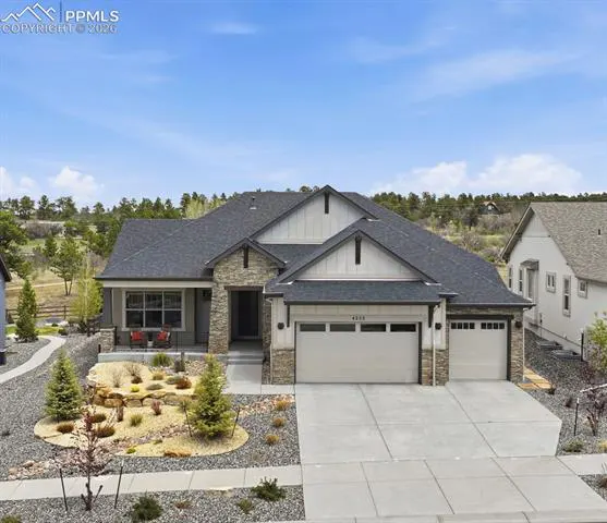 A single-story home with a gray roof, stone accents, and a two-car garage. The front yard has a rock garden and small trees.