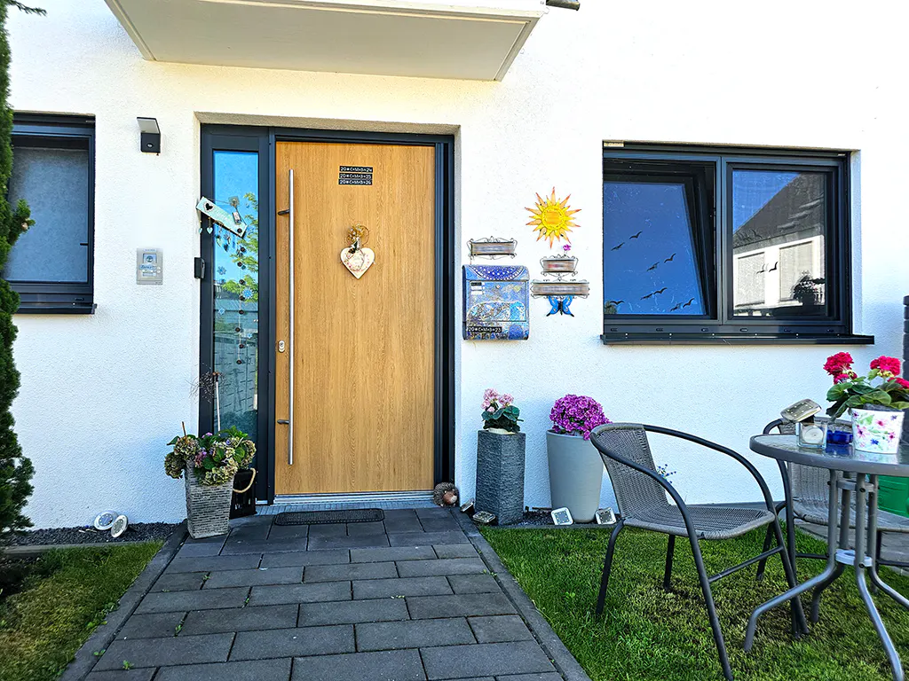 Modern home entrance with a light wood door, stone path, potted flowers, and a small patio set on a green lawn.