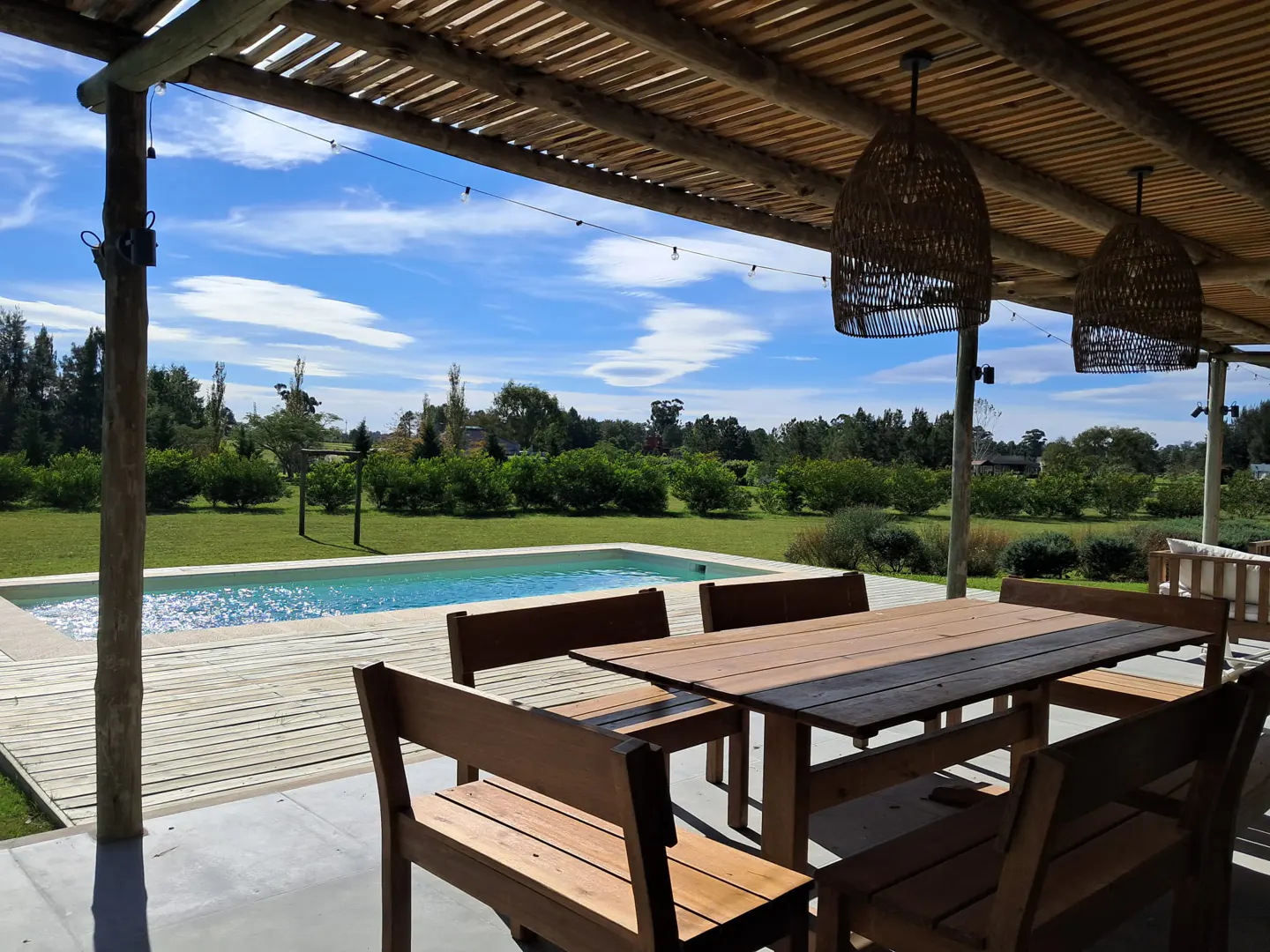 Outdoor dining area with wooden table and chairs under a pergola, overlooking a pool and green lawn. Wicker pendant lights hang from the ceiling.