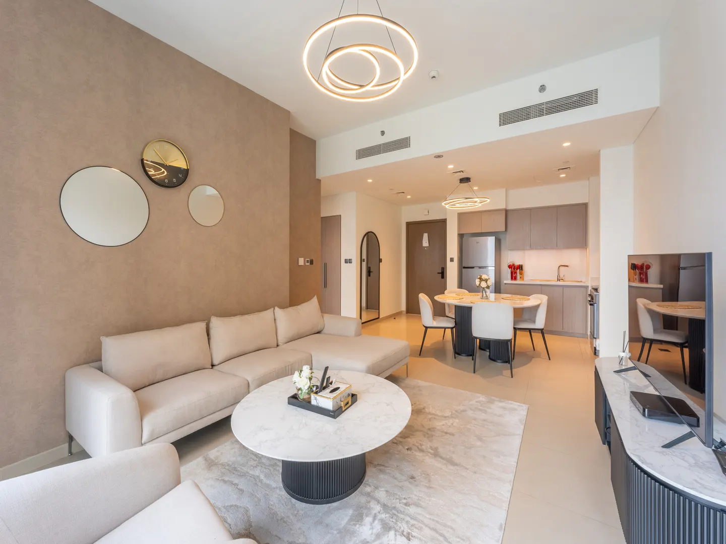 Modern open-plan living room with a beige sectional sofa, marble coffee table, dining area, and kitchen in neutral tones.