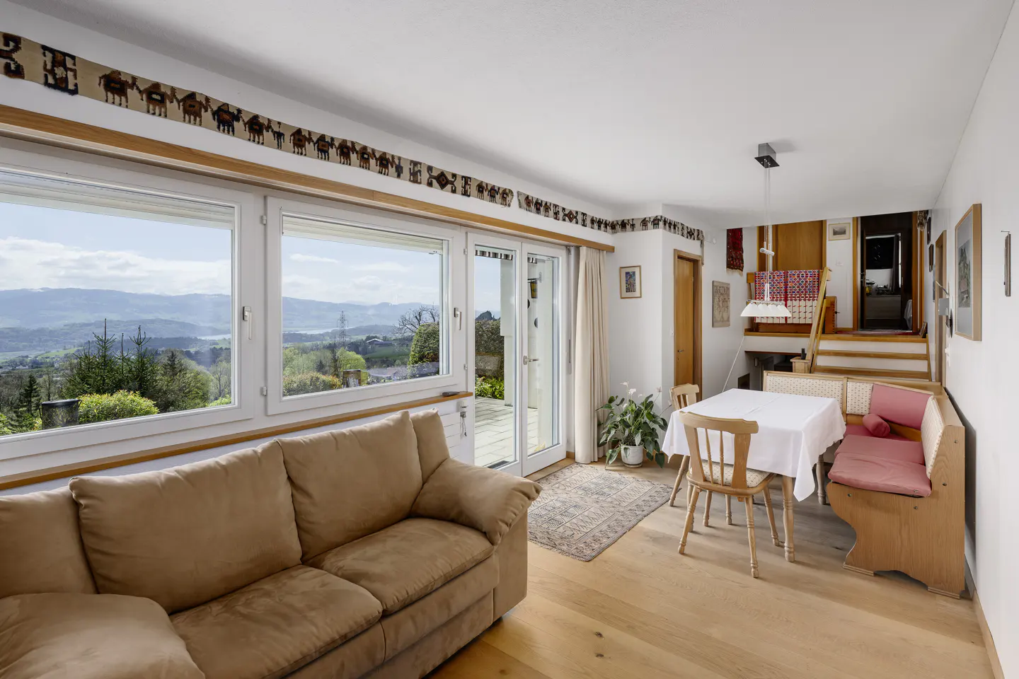 Bright living room with a brown sofa, dining table, and large windows overlooking a green landscape. A decorative border adorns the walls.