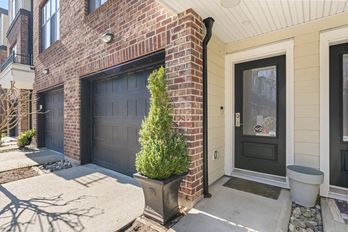 Exterior view of a brick townhouse with a black garage door and front door. A potted evergreen sits next to the garage.