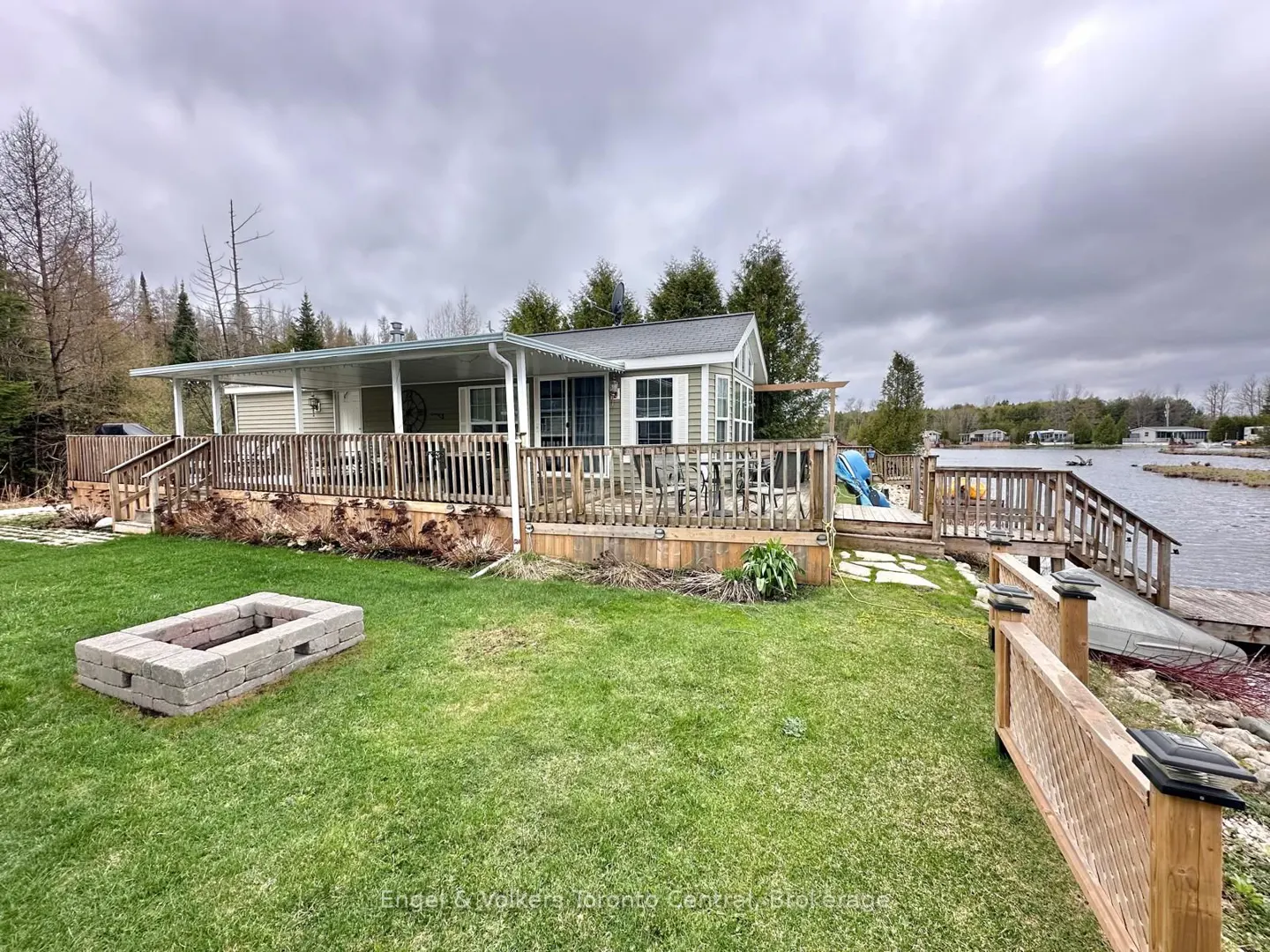 Exterior view of a beige cottage with a wooden deck, fire pit, and lake access on a cloudy day.