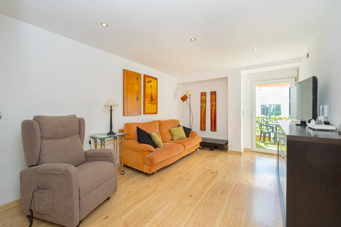 Living room with light wood floors, white walls, and recessed lighting. A brown recliner sits near an orange sofa with decorative pillows. Artwork adorns the walls. A glass door leads to a patio.