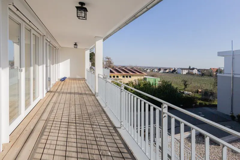 A long, empty balcony with a wooden floor and white railings overlooks a town and vineyard on a sunny day.