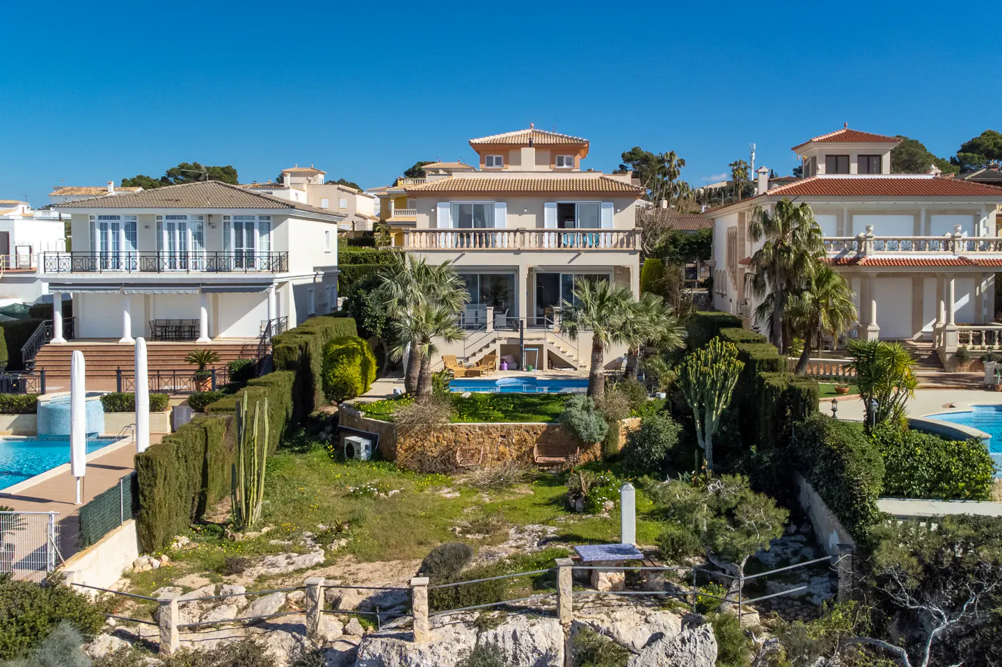 A sunny, elevated view of three Mediterranean-style homes with pools, lush greenery, and a clear blue sky.