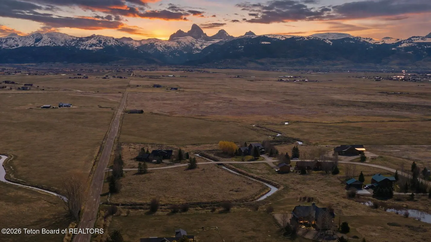 Scenic aerial view of homes in a valley with snow-capped mountains at sunset. The sky is orange and purple.