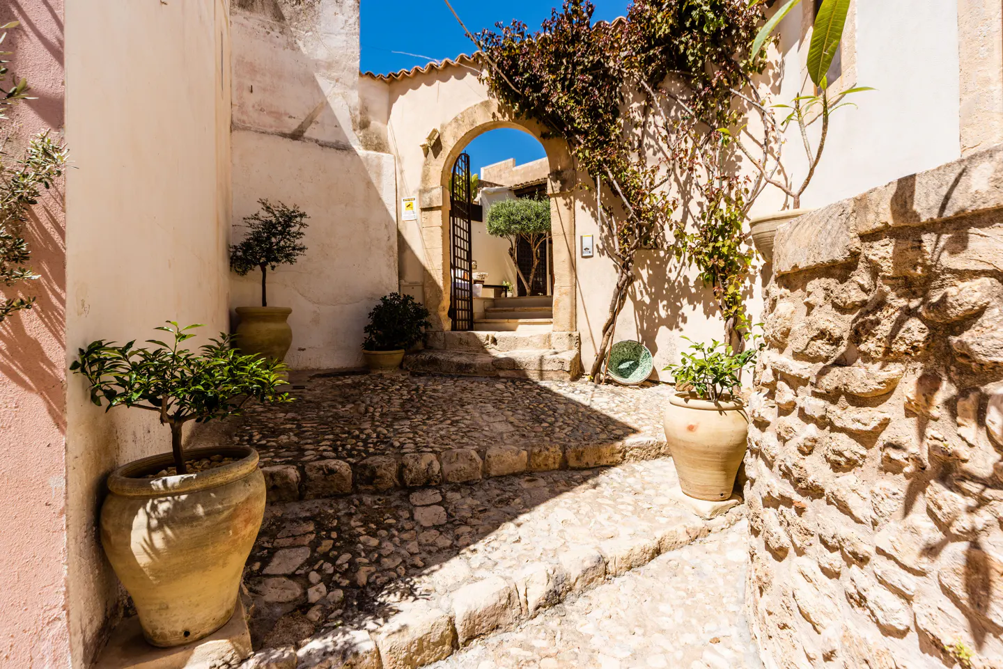 Stone courtyard with potted plants and an arched gateway leading to a garden with a tree.