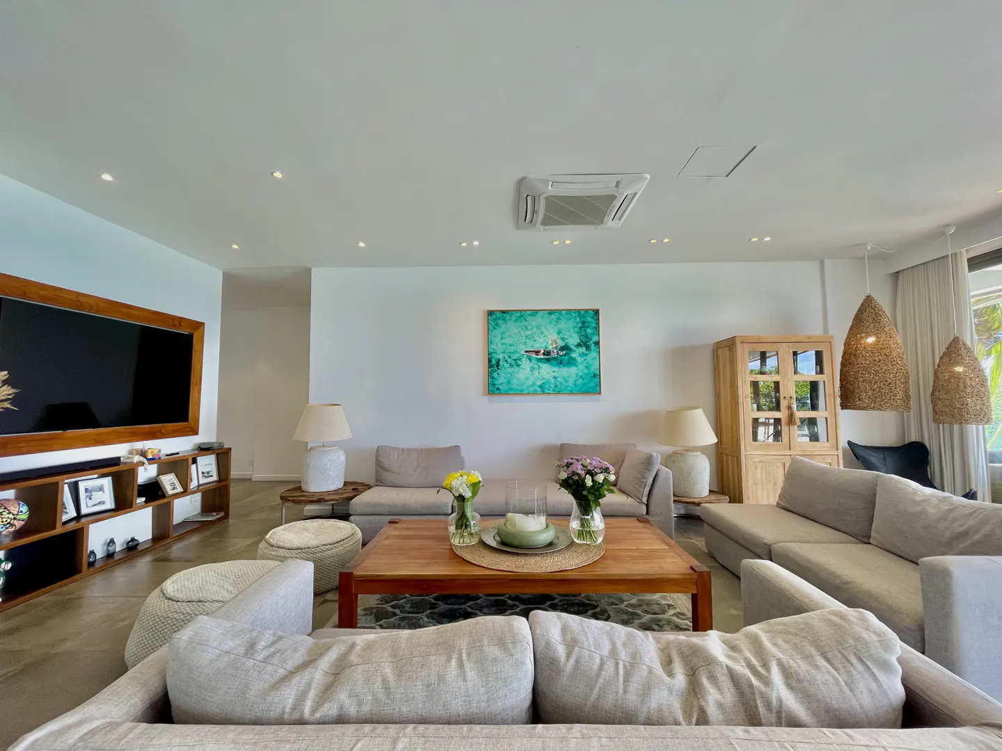 Bright living room with white walls, light gray sofas, and a wooden coffee table with flowers. A large TV and cabinet are on the left.