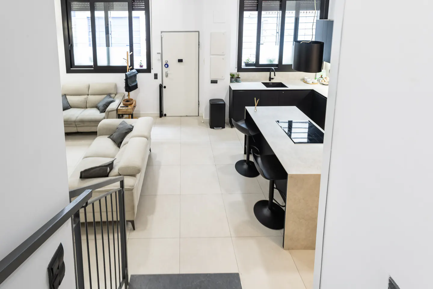 Open-plan living area with light sofas, a kitchen island with black bar stools, and large windows on a tiled floor.