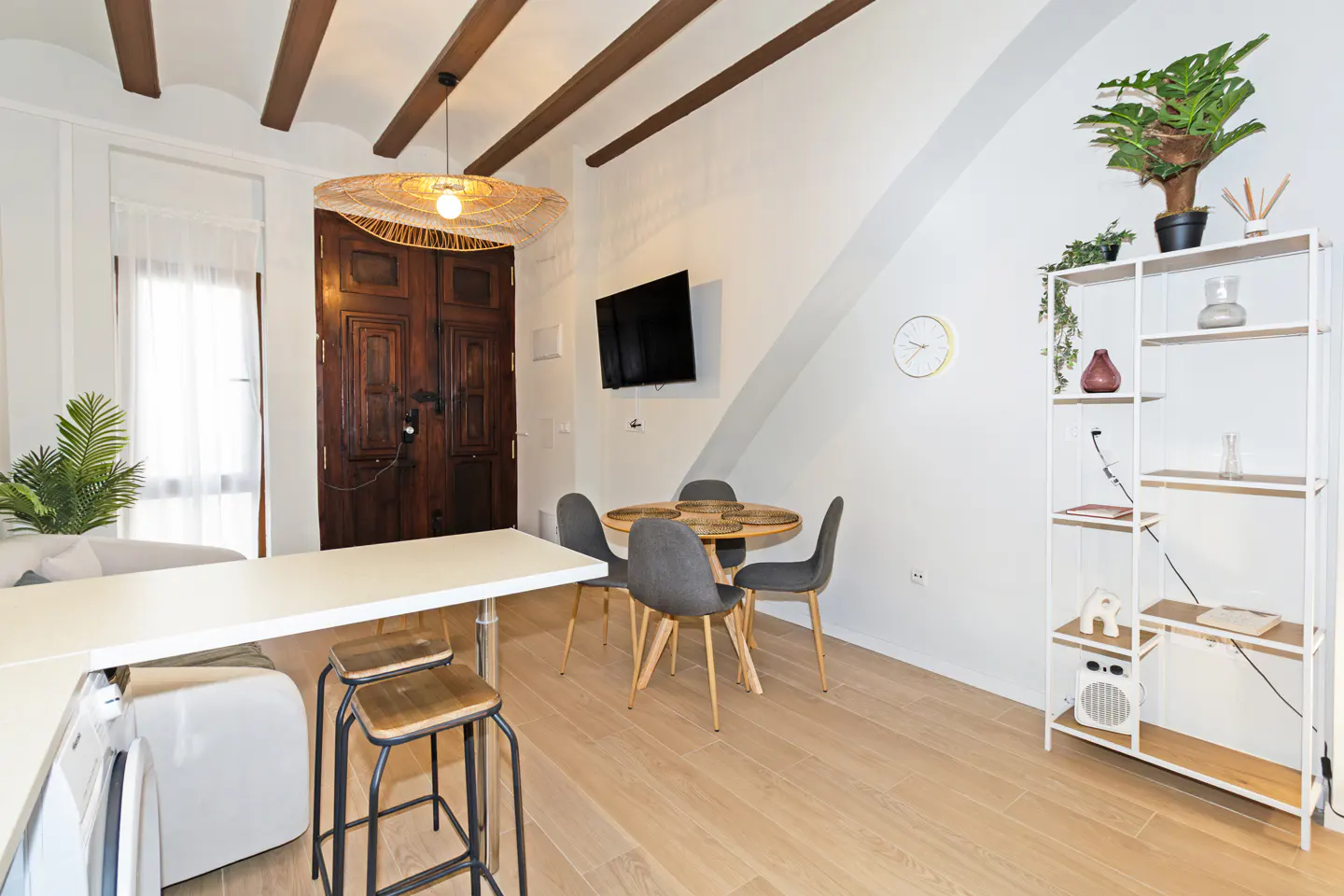 Bright living room with wood floors, white walls, and exposed beams. A round table with four chairs sits near a white bookshelf and a large, dark wood door.