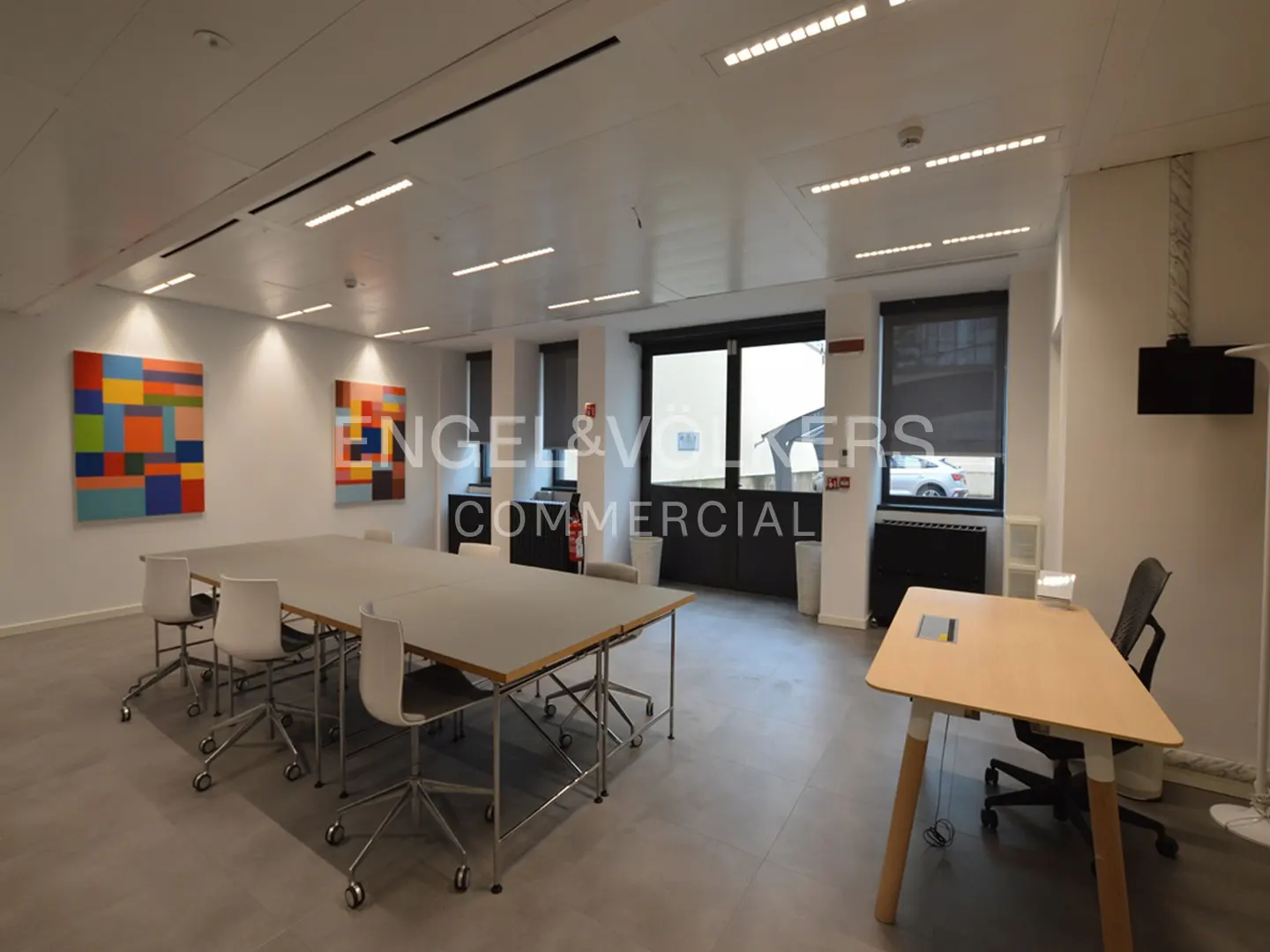 Bright office space with a large conference table, white chairs, and colorful abstract art on the wall. A desk and chair are on the right.