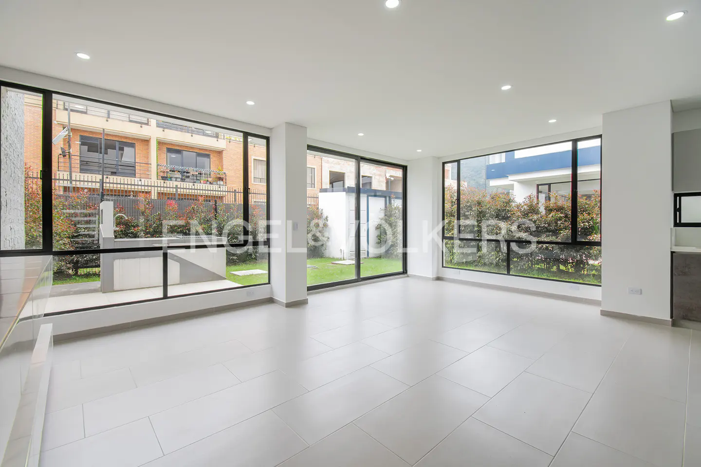 Bright, empty room with white tile floors and large windows. Outside, greenery and buildings are visible.