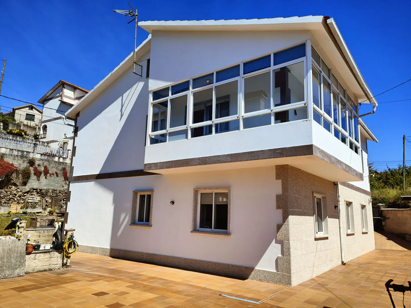 Two-story white house with a glass-enclosed upper balcony, stone corner accents, and a tiled patio under a clear blue sky.