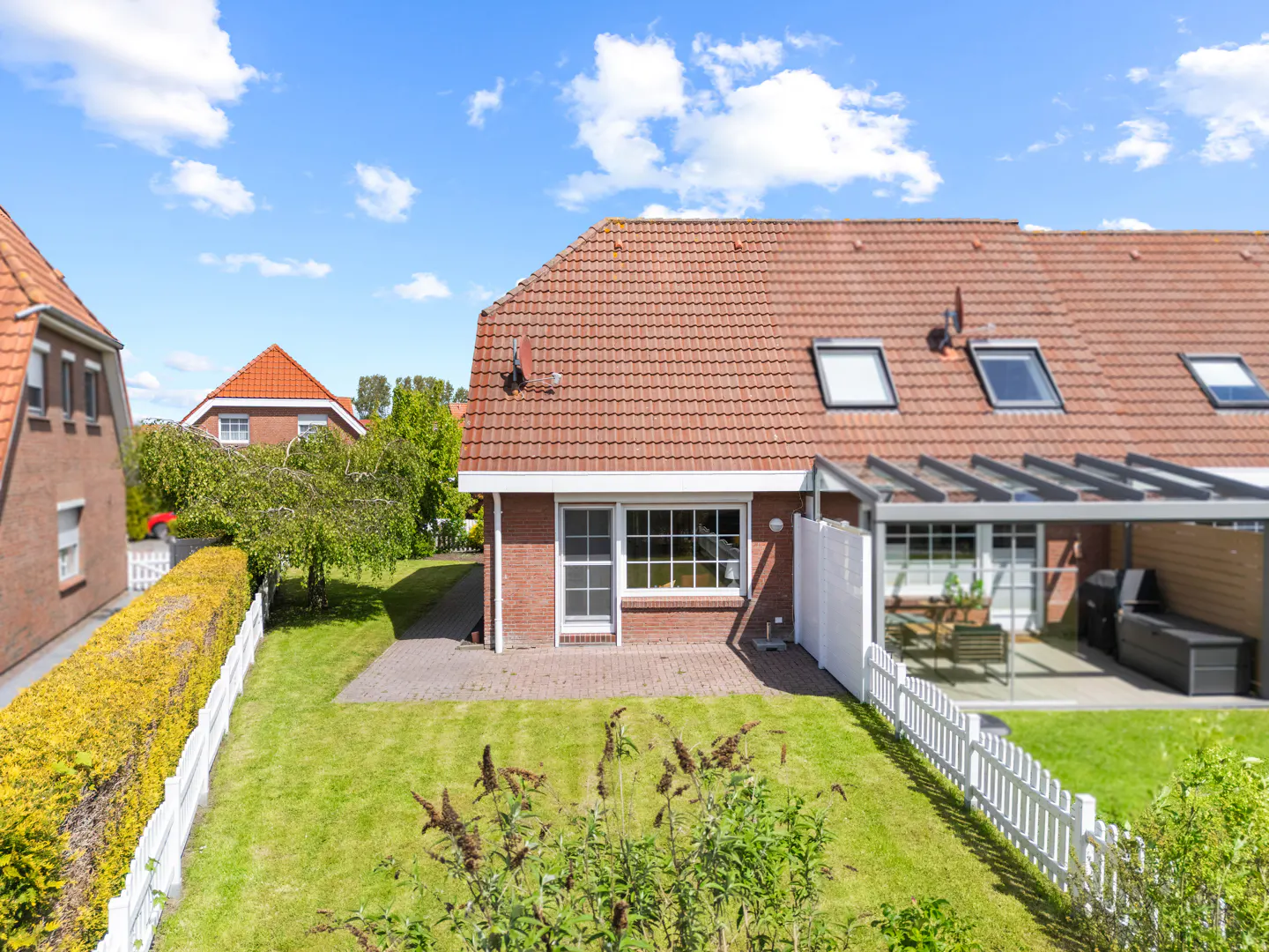 Backyard view of a brick house with a red tile roof, a patio, and a green lawn enclosed by a white picket fence.