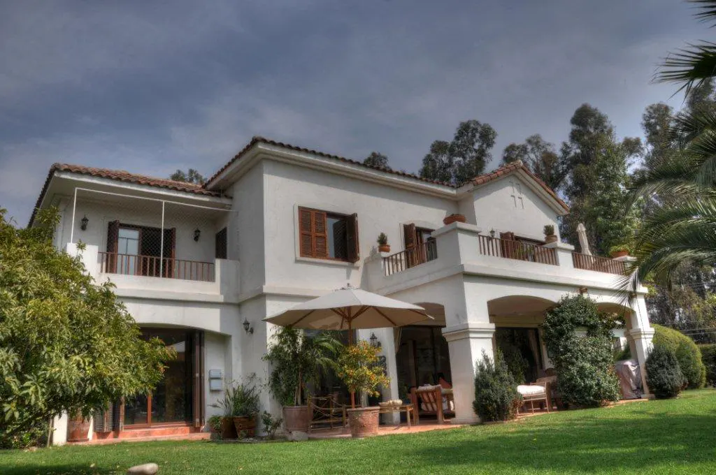 Two-story white house with brown shutters and a red tile roof, surrounded by green lawn and trees. A patio with an umbrella and furniture is visible.