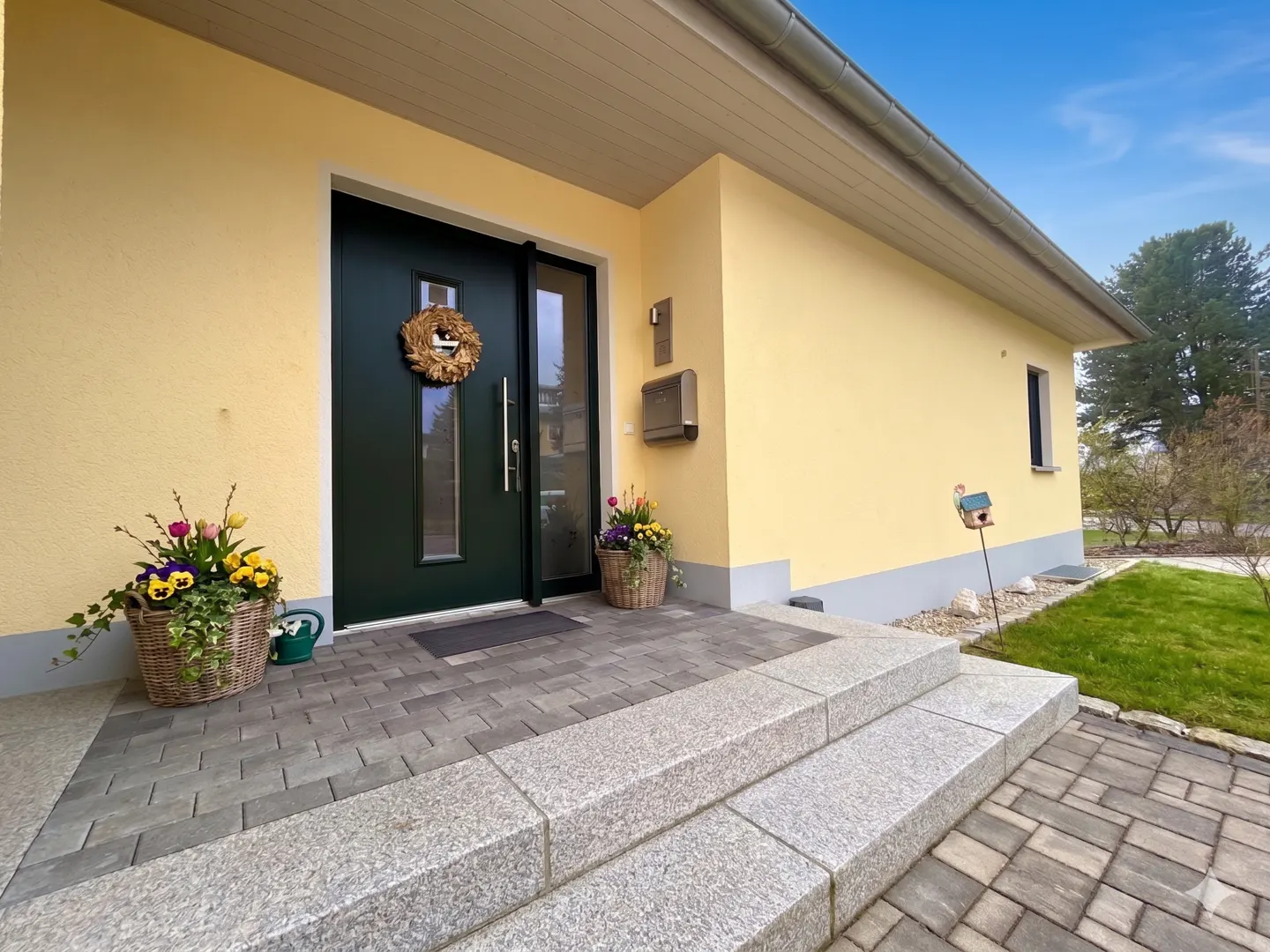 Front of a yellow house with a dark green door, a wreath, and flower baskets on the steps.