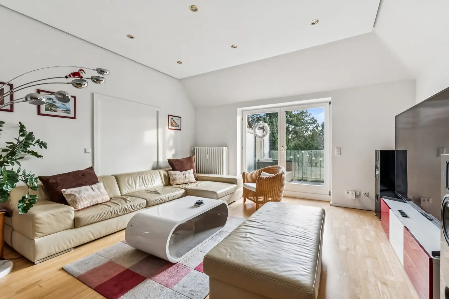 Bright living room with beige leather sofa, white coffee table, and wicker chair. Balcony doors open to trees. Hardwood floors and white walls.
