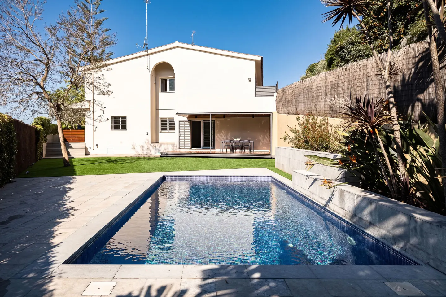 A two-story cream house with a backyard pool, green lawn, and a covered patio area under a clear blue sky.