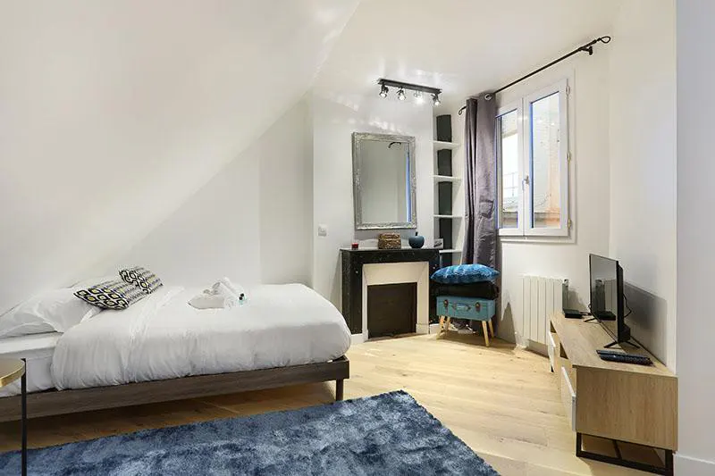 Attic bedroom with white walls, a double bed, a decorative fireplace with a mirror, and a blue rug on light wood floors.