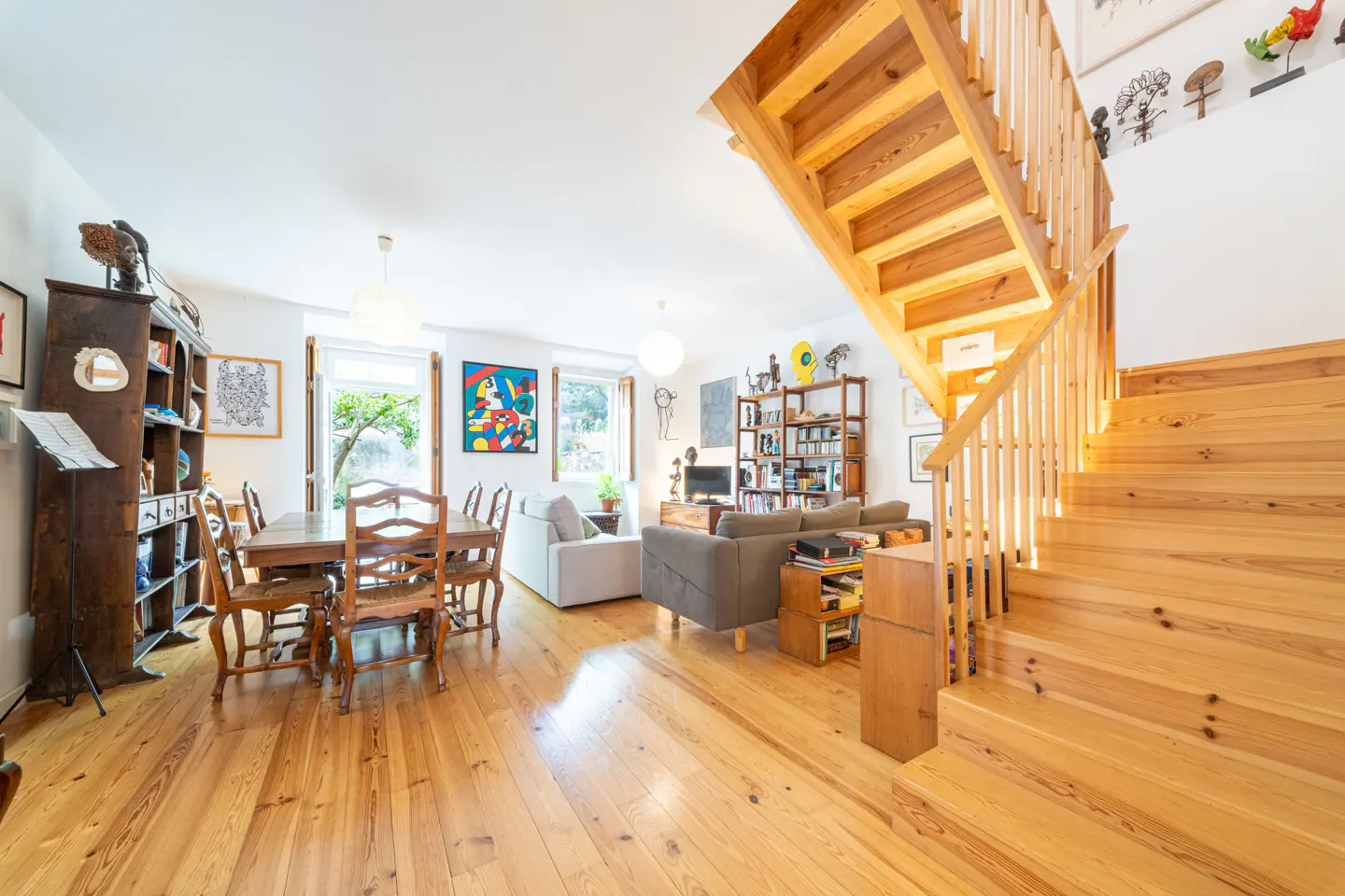 Bright, open-concept living space with wooden floors, stairs, dining table, sofas, and bookshelves. Artwork adorns the white walls.
