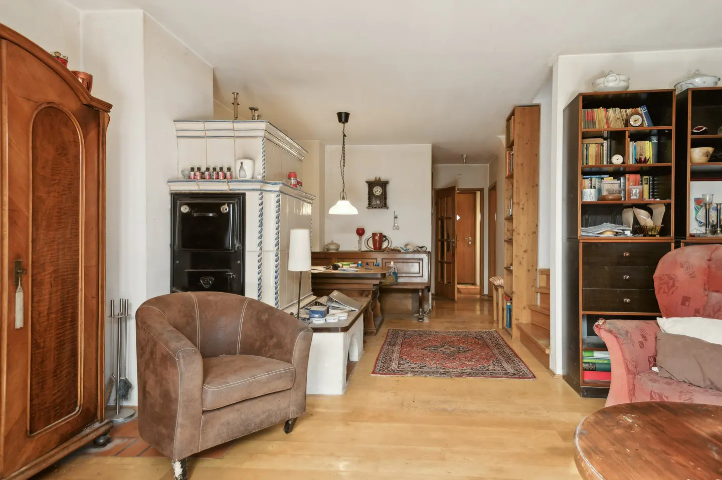 Living room with wood floors, brown leather chair, tiled stove, and wooden cabinet. Bookshelves line the wall.