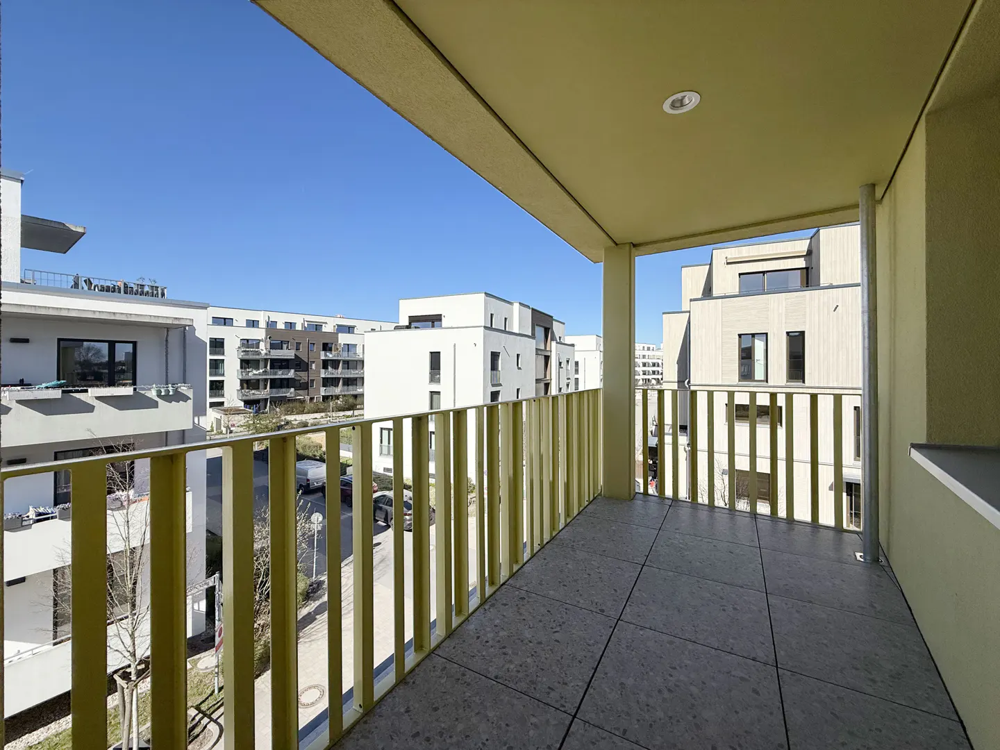 Balcony view with yellow railings and gray tile floor. Modern white buildings are visible in the background under a clear blue sky.