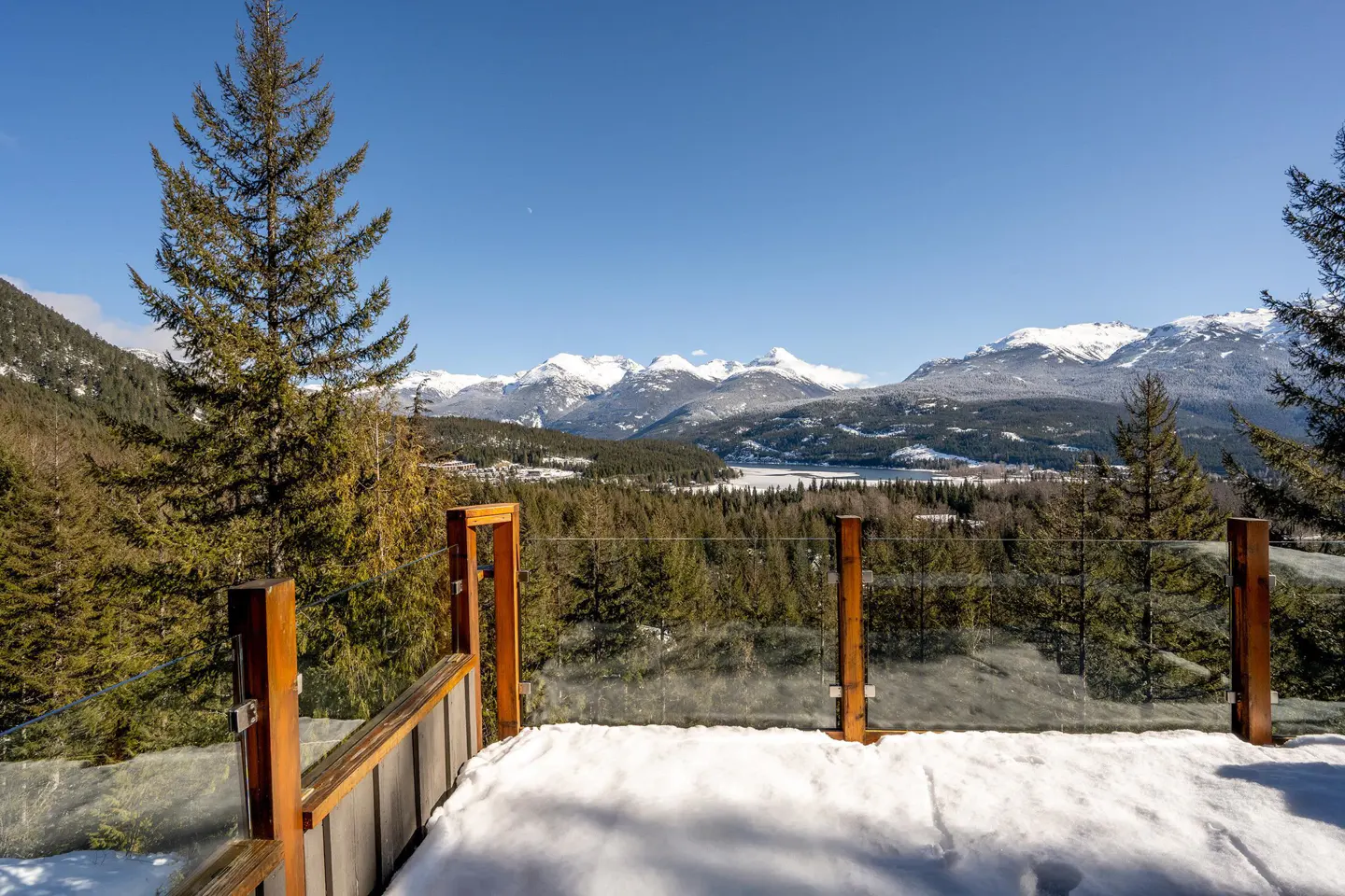 View from a snowy deck with a glass railing overlooking a forest, lake, and snow-capped mountains under a clear blue sky.