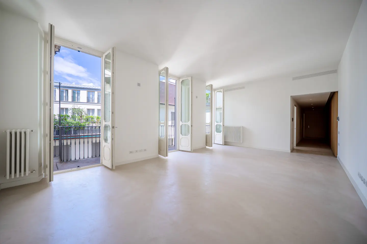 Bright, empty room with white walls, light floor, and three sets of open doors to balconies. Radiators are visible. A dark hallway is on the right.