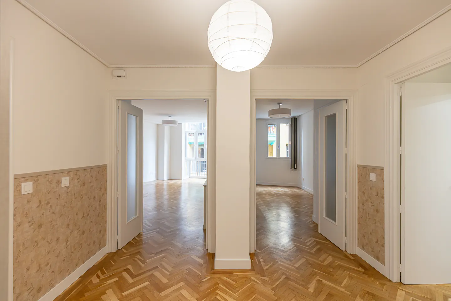 Hallway with herringbone wood floor, white walls, and two open doorways leading to bright rooms. A white paper lantern hangs from the ceiling.