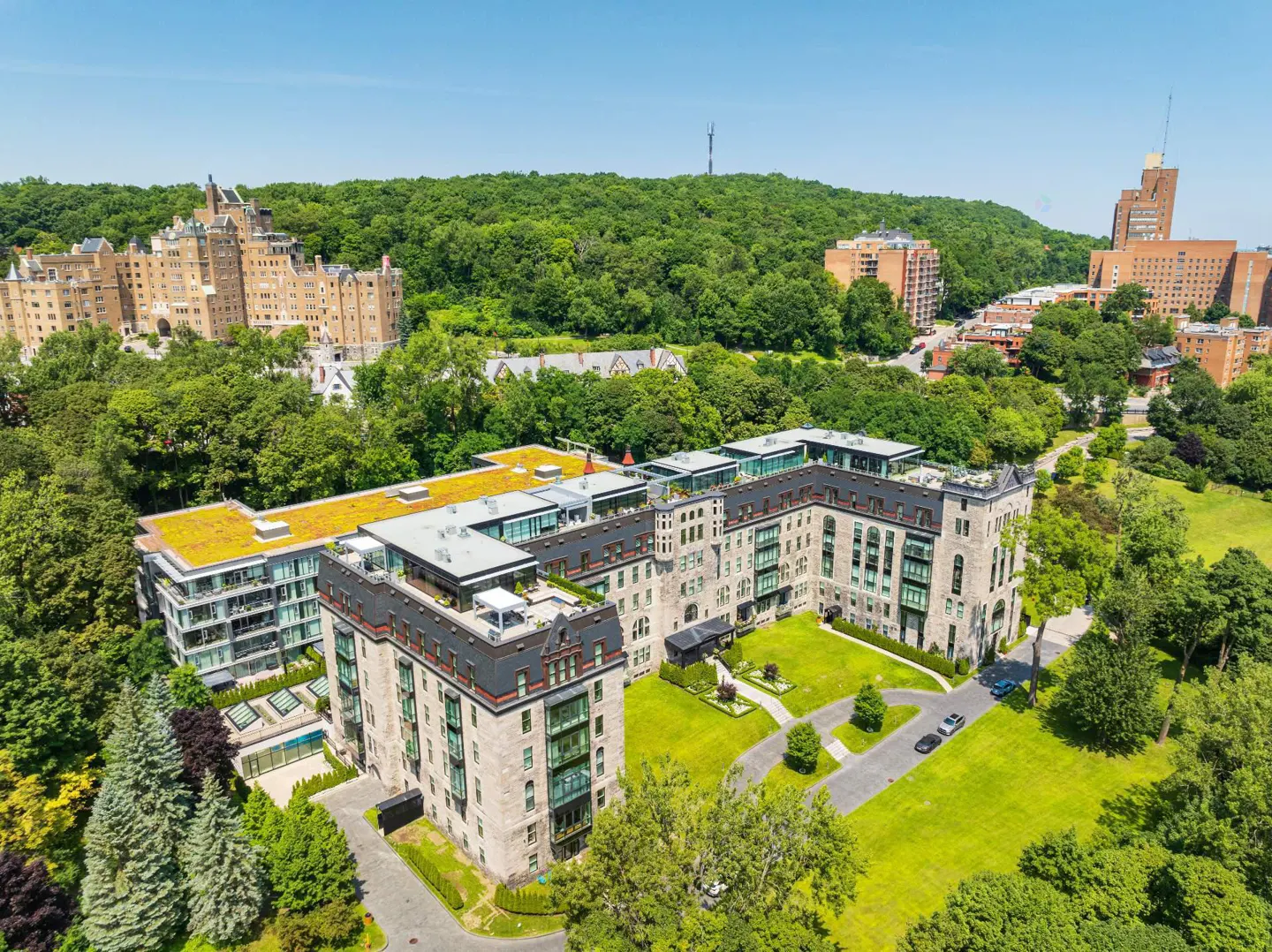 Aerial view of a stone apartment building with green roofs, surrounded by trees and other buildings on a sunny day.