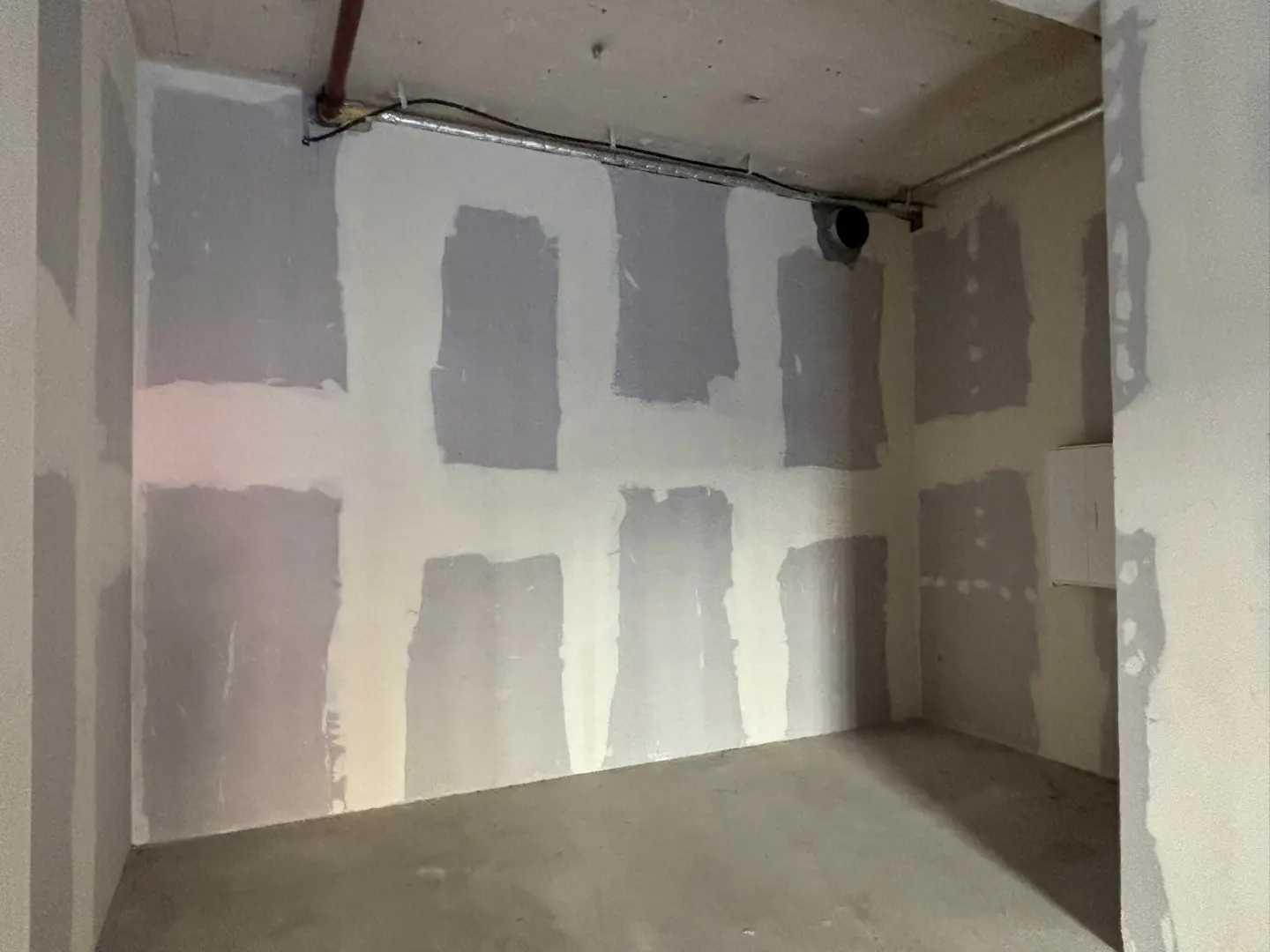 Unfinished room with gray concrete floor and white walls with gray patches. Pipes run along the ceiling.