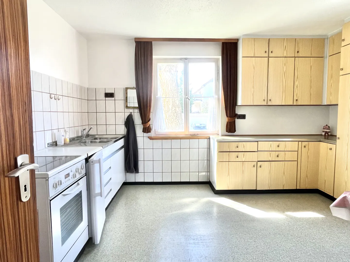 A bright kitchen with white appliances, light wood cabinets, and a window with brown curtains.