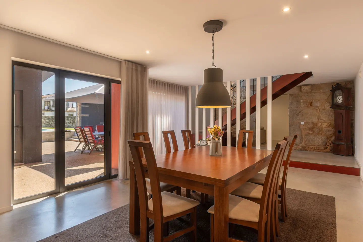 A dining room with a wooden table and chairs, a gray pendant light, and a view of an outdoor patio with furniture.