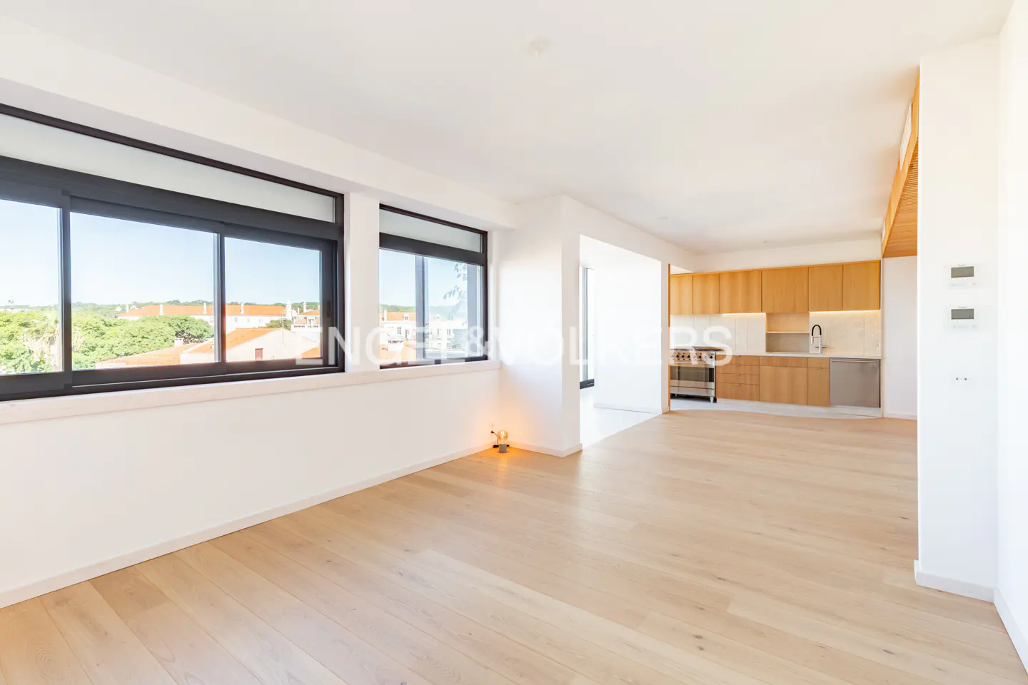 Bright, open-concept apartment with light wood floors, white walls, and black-framed windows overlooking a cityscape. A modern kitchen is visible in the background.