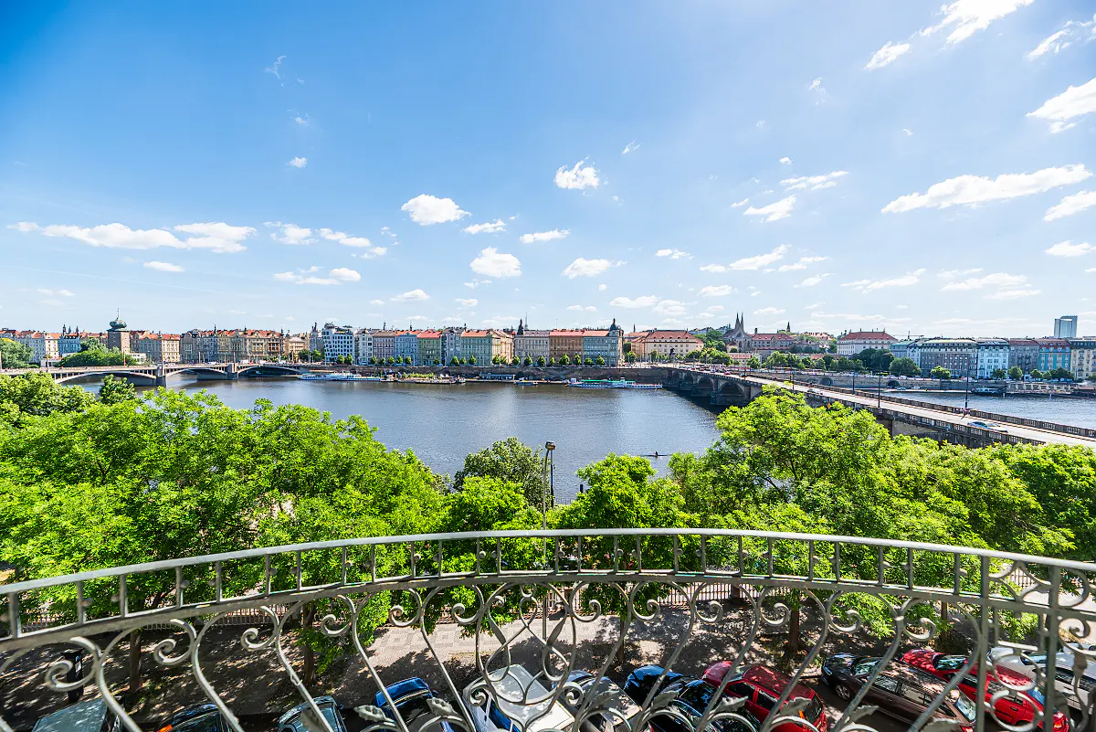 View from a balcony overlooking Prague, Czech Republic, with a river, bridge, and buildings under a blue sky.