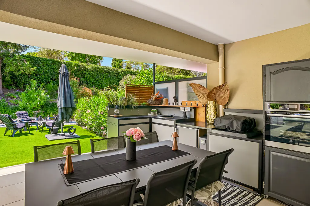 Covered outdoor patio with a black dining table, chairs, and a built-in kitchen with a grill and oven. Green lawn and garden in the background.