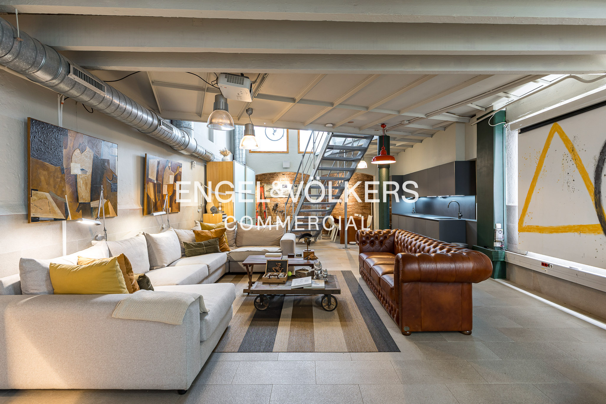 Modern industrial loft living room with a large white sectional, brown leather sofa, metal staircase, and art on walls.