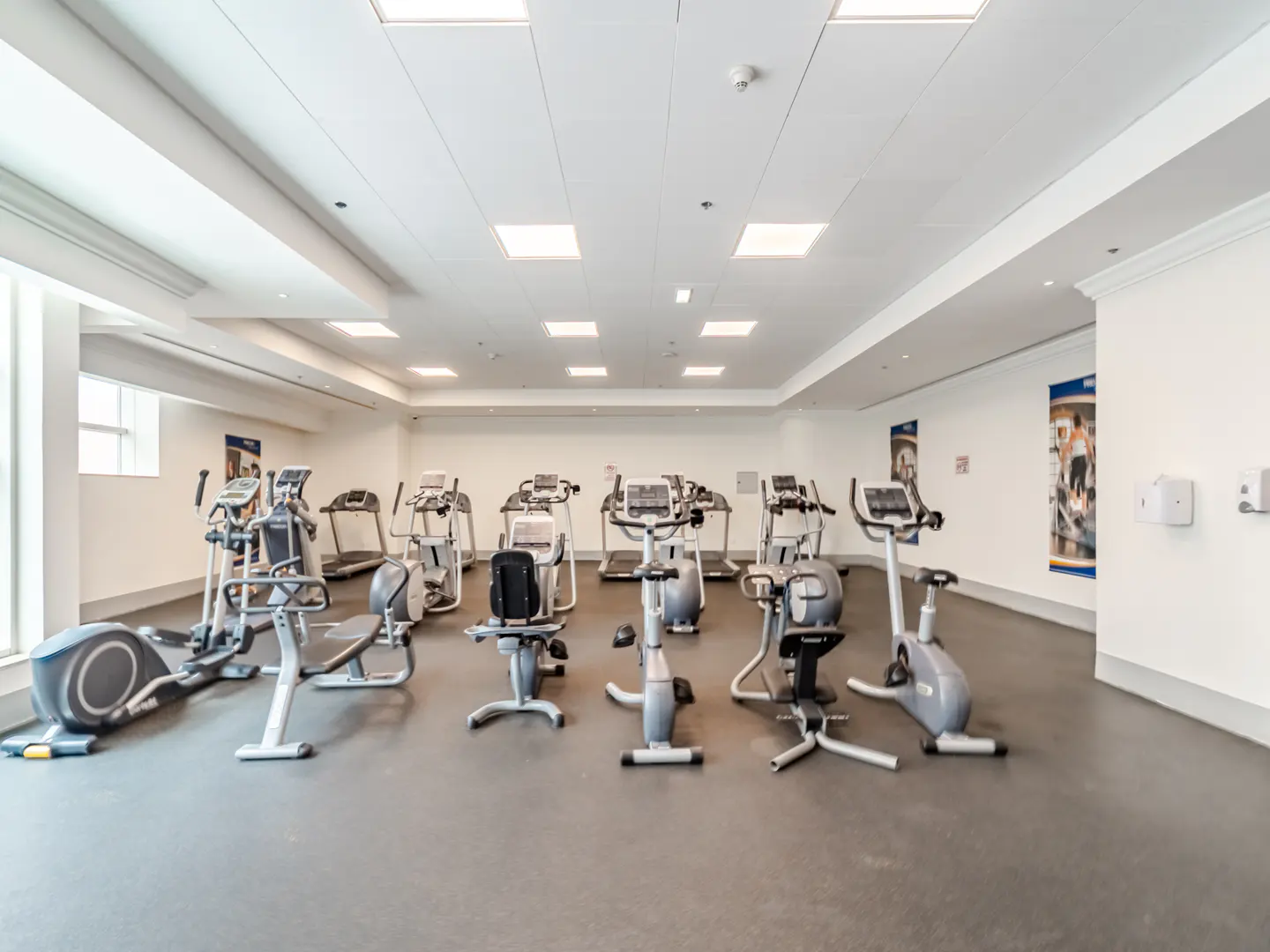 Bright, modern fitness center with rows of grey exercise bikes and elliptical machines on a grey floor under recessed lights.