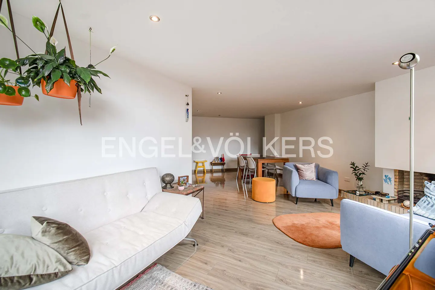 Bright living room with white walls, light wood floors, and modern furniture. Hanging plants, white sofa, blue chairs, and an orange rug add color.