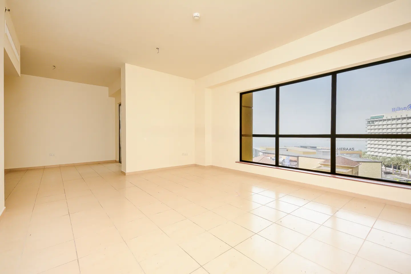 Empty room with beige tile floor, cream walls, and a large window with a view of the Hilton hotel.