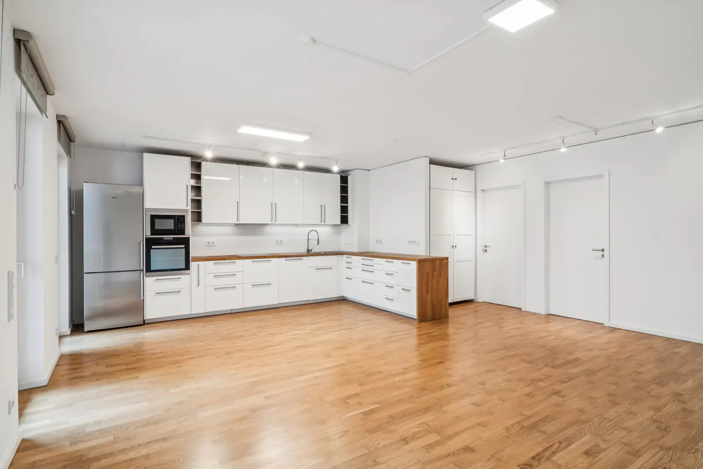 Bright, empty kitchen with white cabinets, stainless steel fridge, and wood floors.