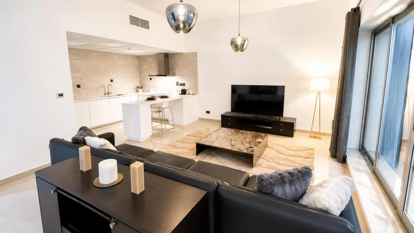 Bright, modern apartment interior with black leather sofa, TV, and white kitchen. Pendant lights hang above a marble coffee table.
